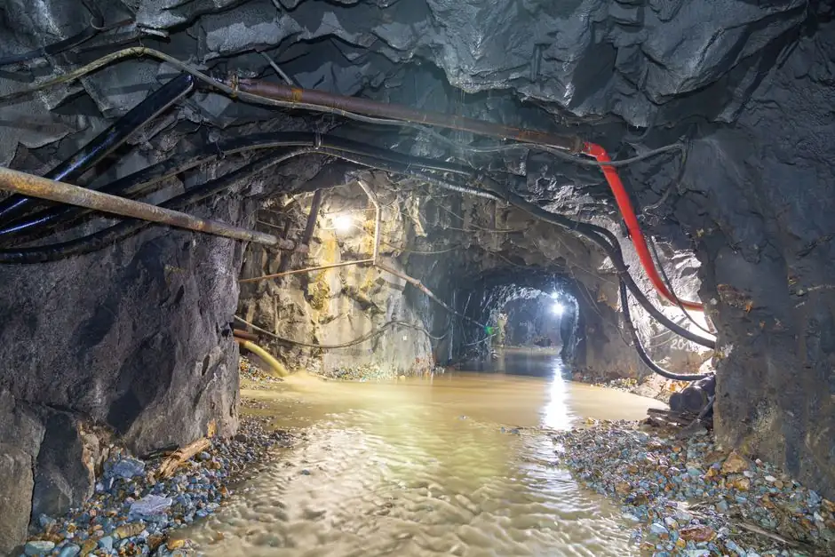 Dimly lit underground cave with flowing water and exposed pipes, showcasing rugged geology.